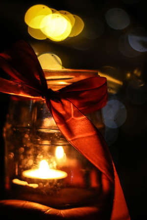 A small candle burns in a jar in the dark against the background of bokeh lights red ribbon bow, bokeh, blurred background, place for textの写真素材