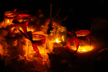A small candle burns in a jar in the dark against the background of bokeh lights red ribbon bow, bokeh, blurred background, place for textの写真素材