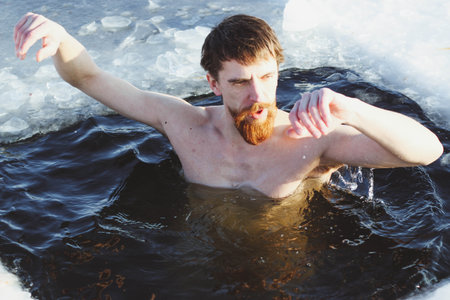 young, slim, handsome, sporty man with a red beard and long hair, naked, in black shorts, diving into ice-cold water in winter, against the backdrop Ukraine, Shostkaの写真素材