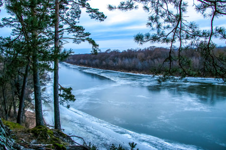 Frozen river covered with snow and ice, top view. Sandy hill with pine trees. Winter forest and...