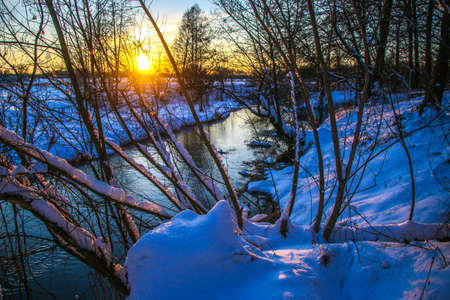 Snow falling on a river with snowy banks tree branches over water, sunny day, the trees are covered with snow Ukraine Shostka, Shostka river Sunsetの写真素材