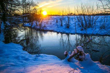 Snow falling on a river with snowy banks tree branches over water, sunny day, the trees are covered with snow Ukraine Shostka, Shostka river Sunsetの写真素材