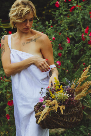 portrait Natural beautiful blonde sexy European girl in a white sundress holds a basket with garden flowers. Walks in the garden on a summer dayの写真素材