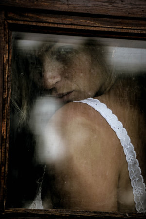 Art portrait of a beautiful young woman looking sadly through an old window in grunge style.