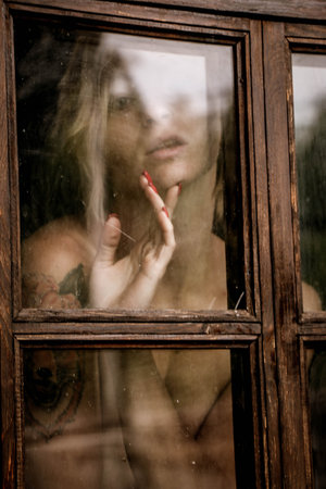 Art portrait of a beautiful young woman looking sadly through an old window in grunge style.