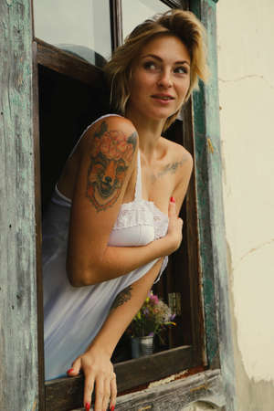 Art portrait of a beautiful young woman, looks through an old window peeps out, sits on the windowsill in grunge style.の写真素材