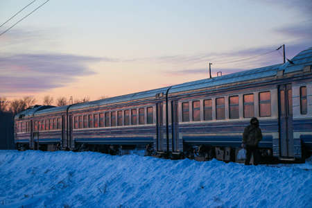 an old passenger train arrives at the station on a cold winter evening at sunset. Snowy landscape, beautiful colored sky.の写真素材