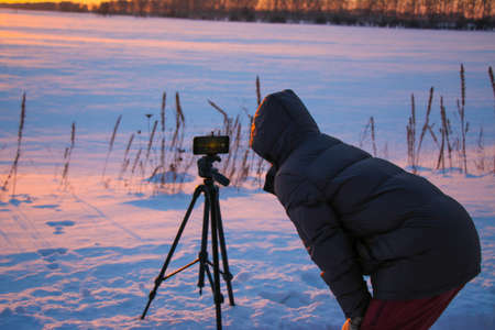 Man with phone on tripod making time-lapse photographs of winter zidio sunset. The field, road and trees are covered with snow.の写真素材