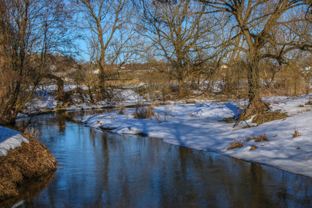 A small river in winter, the sun's rays make their way through the bare trees. Snowy shores are...