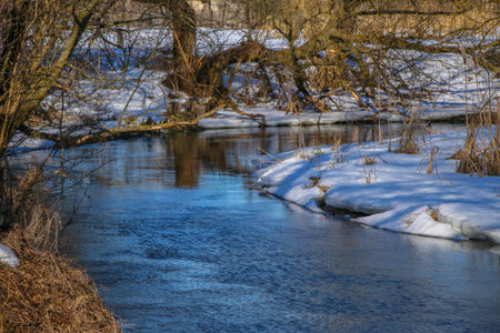 A small river in winter, the sun's rays make their way through the bare trees. Snowy shores are...