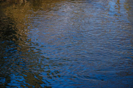Background of trees mirrored on rippled water surface. Small river in winter, reflection of snow-covered banks and sun glare. Natural texture and natural colorの写真素材