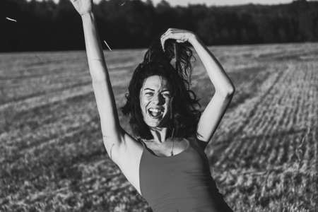 Portrait of a young beautiful brunette in a dress against the background of a mown wheat field, in the summer outdoors, sunset light. Woman happy dancing, black and white photoの写真素材
