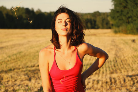 Portrait of a young beautiful brunette in a red dress against the background of a mown wheat field,...