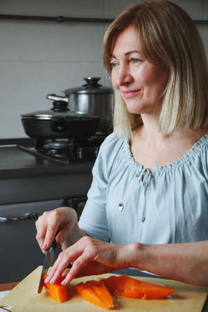 An adult woman, a blonde, calmly prepares food in her home kitchen, cuts a pumpkin in her kitchen, looks to the side or to the camera. A simple household process with real natural light.の写真素材