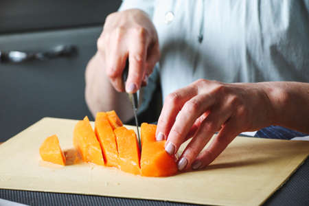 Close-up female adult hands cut orange pumpkin, prepare food in her home kitchen, ordinary household real process natural light, selective sharpness.の写真素材