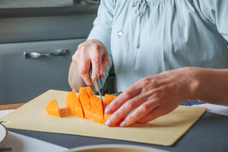 Close-up female adult hands cut orange pumpkin, prepare food in her home kitchen, ordinary household real process natural light, selective sharpness.の写真素材