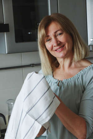 Beautiful adult blonde woman wipes her hands with a towel in her home kitchen, smiles looking at the camera and to the side, natural lightの写真素材