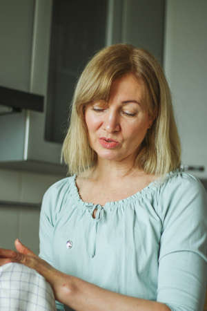 Beautiful adult blonde woman wipes her hands with a towel in her home kitchen, smiles looking at the camera and to the side, natural lightの写真素材