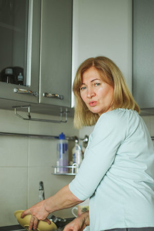 Beautiful adult blonde woman wipes her hands with a towel in her home kitchen, smiles looking at the camera and to the side, natural lightの写真素材