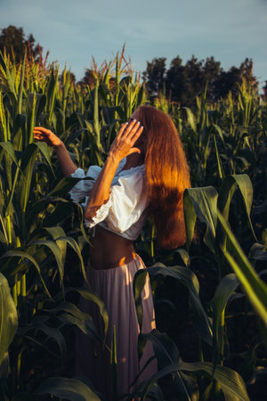 Young beautiful woman with long red hair between green leaves on a corn field in summer, in the light of the setting sun.の写真素材