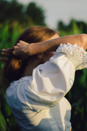 Young beautiful woman with long red hair between green leaves on a corn field in summer, in the light of the setting sun.の写真素材