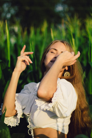 Young beautiful woman with long red hair between green leaves on a corn field in summer, in the light of the setting sun.の写真素材