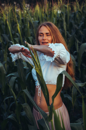 Young beautiful woman with long red hair between green leaves on a corn field in summer, in the light of the setting sun.の写真素材