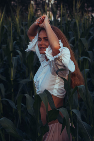 Young beautiful woman with long red hair between green leaves on a corn field in summer, in the...