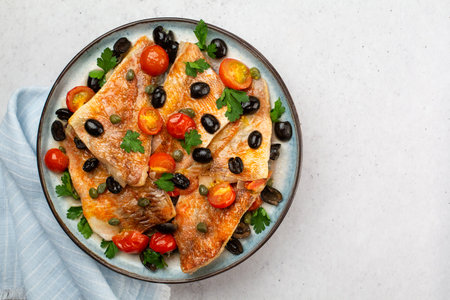 Perch fish fillets stew, italian style, with black olives, tomatoes and capers, with fresh parsley. On light blue plate. On light grey background. Top view.の写真素材
