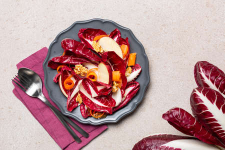 Apple, walnut, carrot and radicchio salad in a dark grey plate. Whole long radicchio. Top view, beige table surface.の写真素材