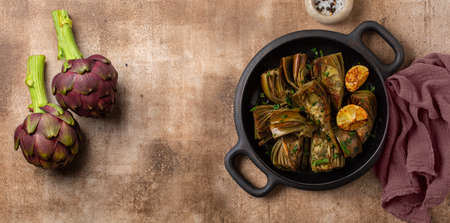 Artichoke or carciofi vegetable, raw and cooked. Artichokes boiled and roasted in olive oil with garlic, garnished with parsley and lemon. Brown  table. Top view. Copy space. Banner size.の写真素材