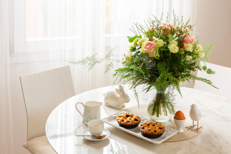 Beautiful morning breakfast table setting, chears. Flowers, espresso coffee, pastry, mini tarts or pies, hard boiled egg. White interior background with window. Spring or easter concept.の写真素材