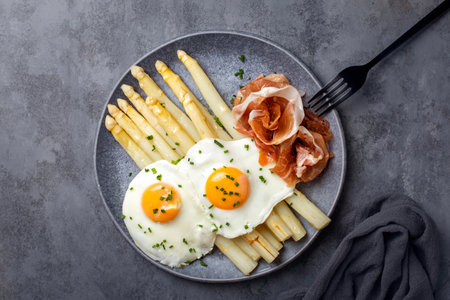 Cooked white asparagus with fried eggs, raw ham or ham, spring onion and butter dressing. The Spruce Eats on a plate. Top view, dark gray background.の写真素材