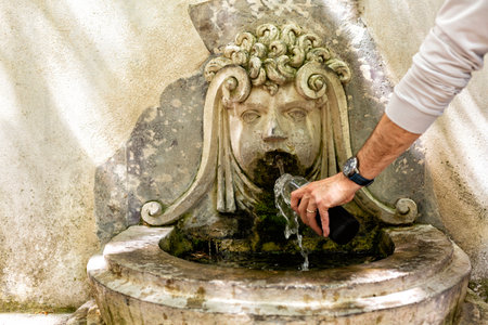 A man filling a glass bottle with drinking water at the beautiful public medieval fountain. Ecological concept.の写真素材