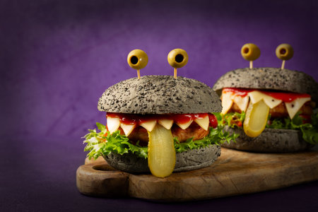 Monster burgers, fun Halloween appetizers, perfect food idea for Halloween party. Made with black charcoal buns, cheese, chicken burger, cheese, letttuce, tomato sauce. Purple background.の写真素材