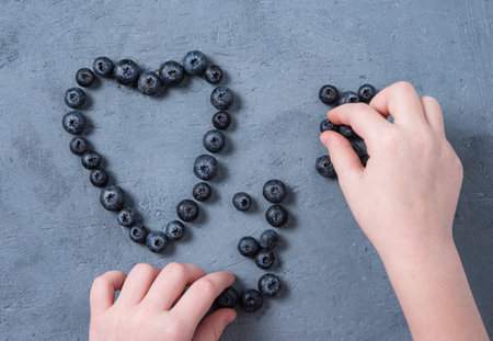 the child's hands collect heart fresh and juicy blueberries on a blue table. Top view anf flat layの写真素材