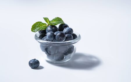 few juicy fresh blueberries in glass bowl with mint leaves on a white background with a hard light. Front view and copy spaceの写真素材