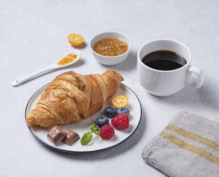 Fresh hot gluten-free croissant on a white plate with kumquat jam, blueberries and raspberries with a Cup of freshly brewed vegan coffee on a white background. Healthy breakfast.の写真素材