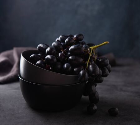 a cluster of juicy black grapes lies in a dark gray bowl on a dark gray table. Photo dark and mood. Front and macro viewの写真素材