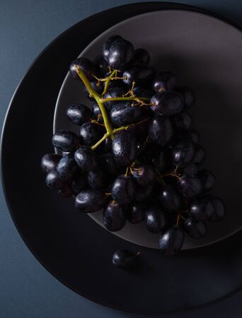 a cluster of juicy black grapes lies in a gray and black plates on a dark gray table. Photo dark and mood. Top viewの写真素材