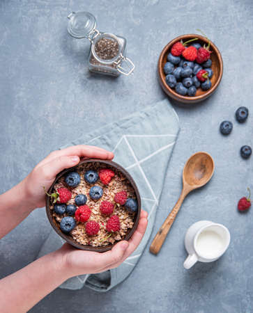 Hands child keep coconut bowl with muesli and berries on blue background. Energy and vegan breakfast. Top viewの写真素材