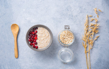 Healthy oats porridge with cranberries on light blue background with jar flakes and oatmeal ears....