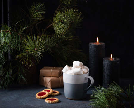 Mug with cocoa, marshmallow and cinnamon on a dark blue background with fruit cookies, lit candles, gifts and fir tree. Dark and mood imageの写真素材