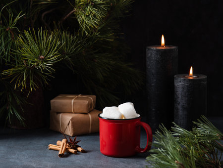 Red mug with cocoa, marshmallow and cinnamon on a dark blue background with lit candles, gifts and fir tree. Dark and mood imageの写真素材