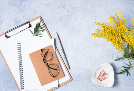 Flat lay with  yellow mimosa  flower,   a glasses, a craft notebook and pencils  on a blue  table....