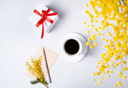 a spring bouquet with yellow mimosa flowers over a cup of black coffee, present box and craft notebook on a white table. concept of 8 March, happy women's day. Top view and copy spaceの写真素材