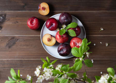 a few juicy red plums in blue plate with branch blossom flowers on a napkin on a brown wood background. Top view and copy  space  imageの写真素材