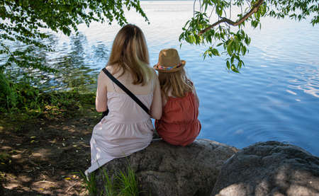 two girls are sitting on a rock under a tree on the shore of a lake on a sunny summer  day. Without faceの写真素材