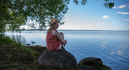 a little girl in an orange dress and hat is sitting on a stone on the shore of a lake  and looking dreamily into the distance on  a summer dayの写真素材