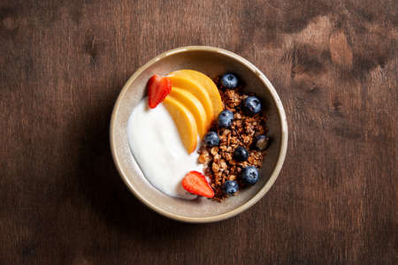 Fresh natural yogurt with homemade granola, peach, strawberry and blueberry in a bowl on a dark wooden background. The concept healthy and nutritious breakfast. Diet food. Top view and copy space.の写真素材